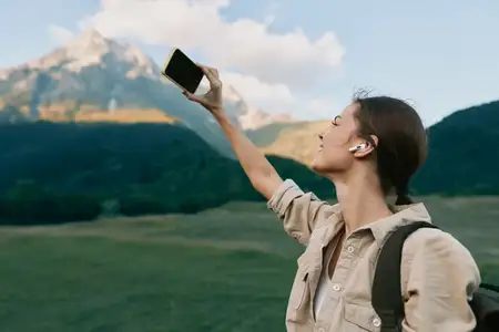 Молодая женщина в наушниках и бежевой рубашке поднимает смартфон, чтобы сделать селфи на фоне гор и зеленых полей под ясным голубым небом