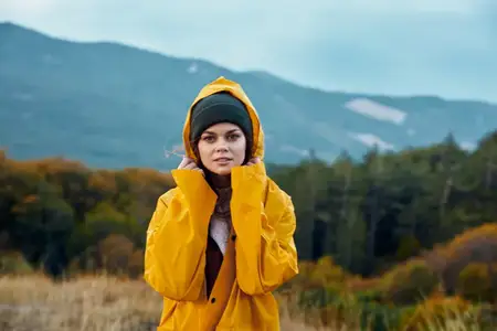 Женщина любуется живописной красотой горного пейзажа, стоя в жёлтом дождевике в поле во время туристической экспедиции