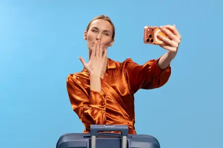 Молодая женщина в оранжевой атласной рубашке посылает воздушный поцелуй в камеру, одновременно делая селфи на смартфон