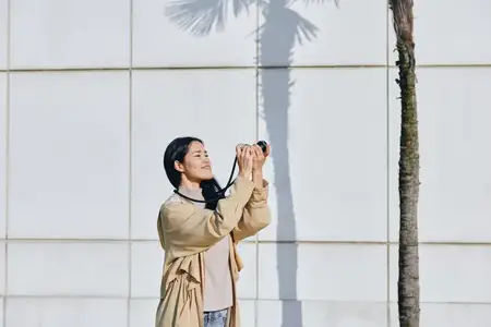 Уличная сцена: женщина-фотограф с камерой рядом с тенью от пальмы и плиточной стеной