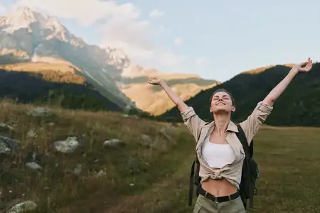 Женщина-любитель активного отдыха стоит на открытом поле с горами на заднем плане, улыбается с поднятыми руками, запечатлевая яркий момент образа жизни, полный исследований, хорошего самочувствия и…
