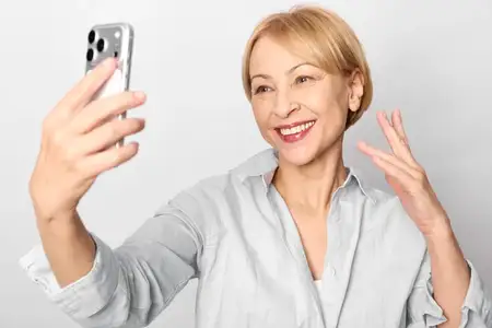 Селфи: улыбающаяся пожилая женщина с телефоном и жестом приветствия