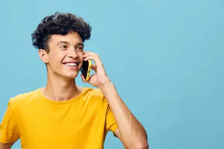 Улыбающийся молодой человек разговаривает по телефону, одетый в ярко-желтую футболку, на фоне успокаивающего синего фона, что отражает чувство позитива и связи