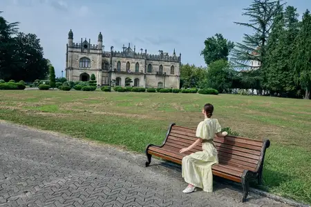 Женщина в бледном платье отдыхает на скамейке в парке, любуясь архитектурой старинного замка, в то время как деревья обрамляют пейзаж