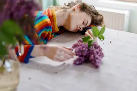 Непринужденная сцена: женщина дома пишет за столом, рядом стоят цветы сирени