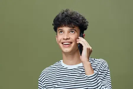 Улыбающийся молодой человек в полосатой рубашке разговаривает по смартфону, смотрит вверх с радостью и увлеченностью, простой оливково-зеленый фон, естественное освещение, тема общения