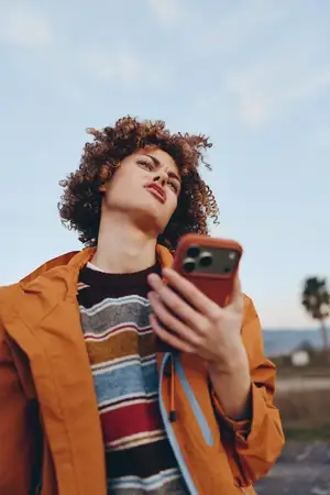 Женщина с кудрявыми волосами поднимает взгляд, держа смартфон для селфи на улице под мягким солнечным светом