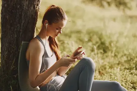 Молодая женщина в спортивном наряде взаимодействует со смартфоном, слушая музыку, сидя у дерева в мирной обстановке на открытом воздухе