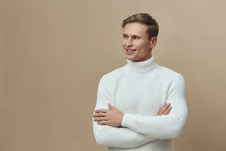 Счастливый мужчина в белом свитере с высоким воротником улыбается и смотрит в сторону в студийной обстановке на бежевом фоне