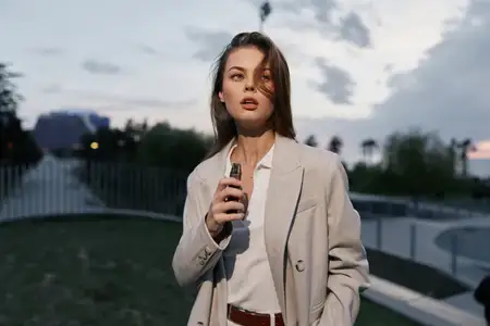 Молодая женщина европейской внешности вейпит на открытом воздухе в сумерках, одетая в бежевый пиджак и белую блузку, задумчивое настроение, городская обстановка с мягким вечерним светом…