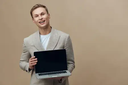 Улыбающийся мужчина держит ноутбук с пустым экраном, одет в бежевую куртку и белую футболку, стоит изолированно на бежевом фоне с уверенным выражением лица