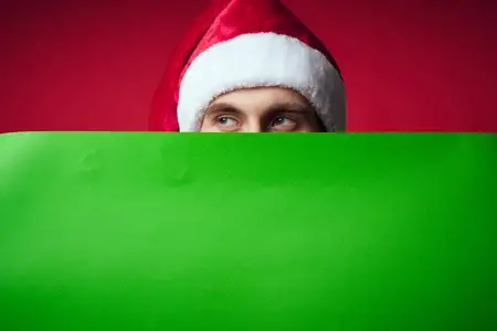 Эмоциональный мужчина в рождественской шапке с зелёным макетом, позирующий в студии