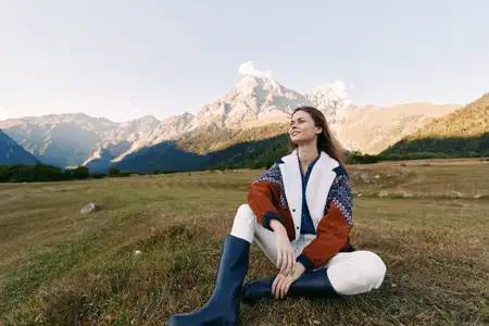 Женщина в осеннем наряде позирует на травянистом лугу на открытом воздухе с видом на горы, в сапогах и уютной куртке