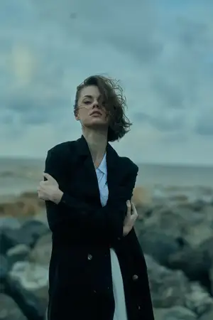 Уличная модная сцена с ветром, пальто и моделью на ветреном побережье, скалистый фон, мрачная атмосфера, сильная поза и стильное отношение, запечатленные при естественном освещении