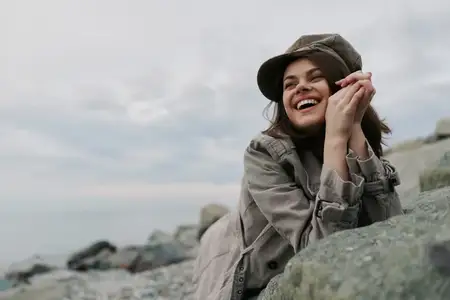 Счастливая молодая женщина в теплом пальто и шляпе опирается на камни у моря