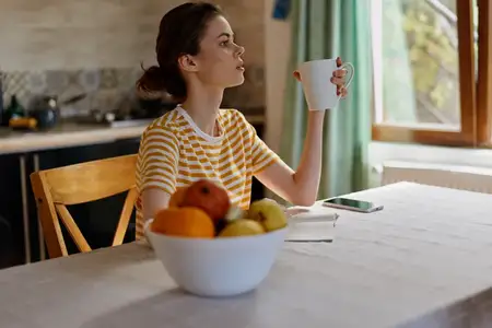 Жизненный портрет женщины дома: утренняя рутина с кофе, чаша с фруктами, телефон рядом, тёплая кухонная обстановка, расслабленное выражение лица, естественный жизненный снимок, уютный декор…
