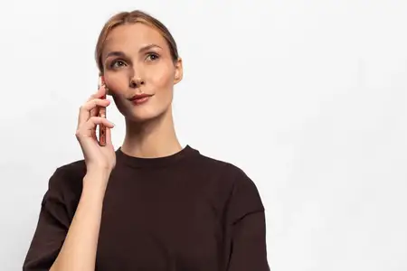 Задумчивая молодая женщина в коричневой рубашке прижимает смартфон к уху, глядя в сторону на белом фоне