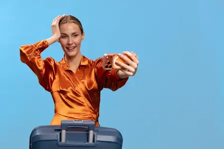 Улыбающаяся молодая женщина в оранжевой блузке делает селфи с чемоданом на голубом фоне