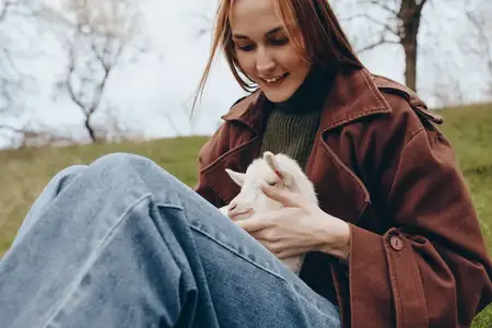 Молодая женщина в коричневой куртке и синих джинсах сидит на траве и нежно держит на руках маленького белого козленка, наслаждаясь мирным моментом на природе