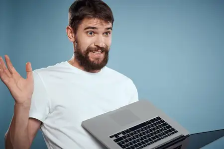 Эмоциональный мужчина с ноутбуком в руках на синем фоне