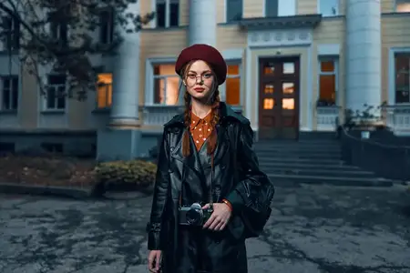 Модная женщина в красном берете и кожаной куртке позирует на фоне городского здания, излучая уверенность и стиль