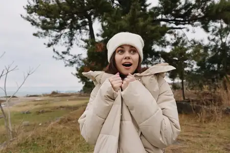Сцена на открытом воздухе: модная женщина в тёплом пальто и шапке-бини, удивлённая чем-то за кадром, на фоне серого неба и природного ландшафта в ветреный день