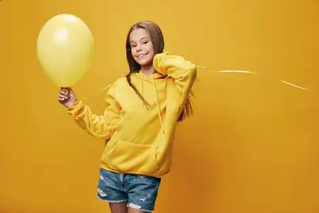 Юная девушка в джинсовых шортах и желтой толстовке уверенно улыбается, держа за нитку ярко-желтый воздушный шар