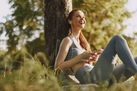 Молодая женщина в спортивной одежде улыбается, слушая музыку, сидя под деревом, что олицетворяет расслабление и радость жизни на открытом воздухе