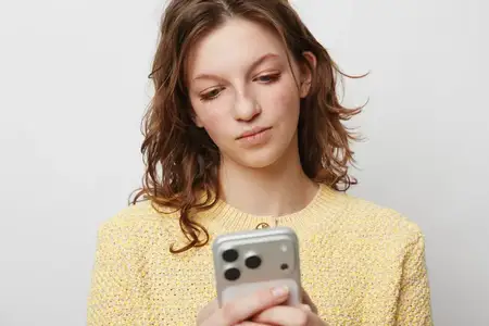 Женщина с вьющимися волосами в желтом вязаном свитере держит смартфон и смотрит на экран со спокойным лицом