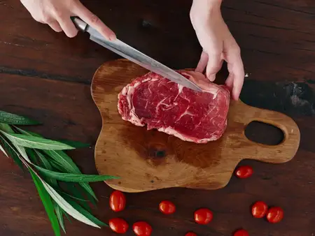 Женщина с ножом в руке нарезает свежее мясо стейка для жарки на кухне; на столе соль, перец и другие специи, красные помидоры черри и зелень; приготовление ужина в домашней кухне