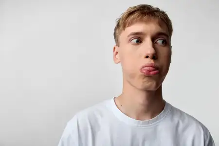 Озорной высунутый язык, выразительный молодой человек в футболке, игривая и юмористическая поза, крупный план деталей лица с легким студийным свечением на однотонном фоне, непринужденная…