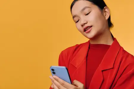 Молодая женщина в красной рубашке и куртке держит смартфон с закрытыми глазами, изолированная на жёлтом фоне, что символизирует цифровое взаимодействие и использование телефона