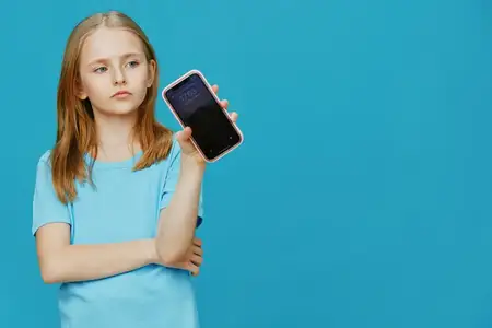Задумчивая девочка в голубой рубашке держит смартфон, демонстрируя любопытство к технологиям на фоне ярко-голубого фона