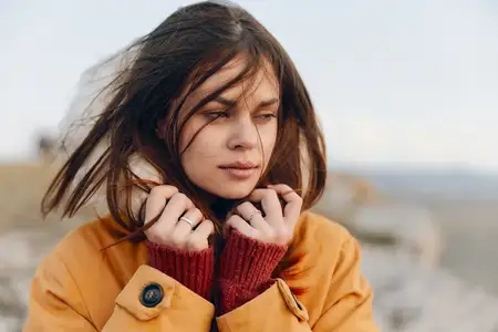 Молодая женщина в оранжевой куртке стоит на вершине холма, закрыв лицо руками, созерцая красоту и безмятежность природы