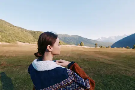 Женщина в узорчатой куртке и флисовом пальто смотрит через травянистый луг в сторону гор, профиль на открытом воздухе