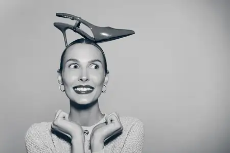 Счастливая светлокожая женщина-европеоид в серьгах балансирует одной туфлей на высоком каблуке на голове и смотрит в сторону в студийном портрете