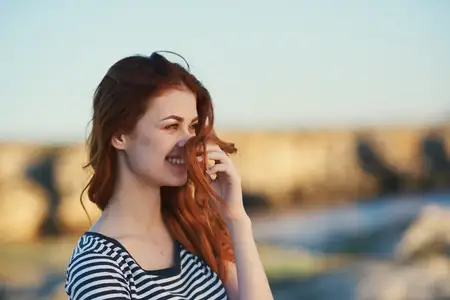 Счастливая женщина в полосатой футболке, с рыжими волосами, улыбается и позирует как модель