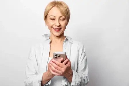 Улыбающаяся пожилая женщина пользуется смартфоном, смотрит на экран с теплым выражением лица