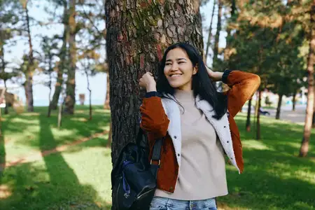 Улыбающаяся женщина в парке, прислонившаяся к дереву, в многослойном кардигане и свитере, с рюкзаком, готовым к прогулке, солнечный день, спокойная уличная сцена, подчеркивающая природу и…