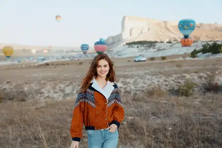 Женщина в джинсах и оранжевой куртке стоит перед полем, на фоне которого видны воздушные шары