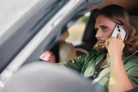 Мужчина в зеленой рубашке сидит в автомобиле, прижав смартфон к уху, погруженный в размышления