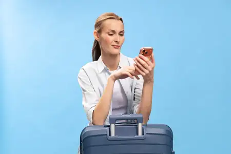 Привлекательная молодая женщина в повседневной белой рубашке держит смартфон, стоя за синим чемоданом на голубом фоне