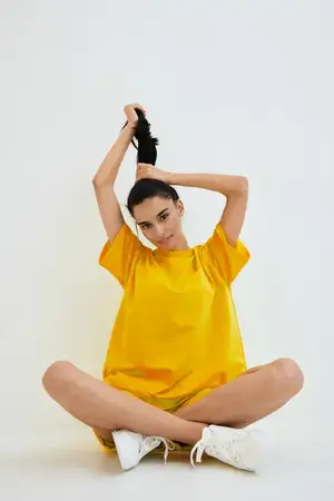 Молодая женщина в желтой oversized рубашке сидит, скрестив ноги, на белом полу и завязывает свои длинные темные волосы, на ней белые кроссовки, минималистичная светлая обстановка, повседневное…