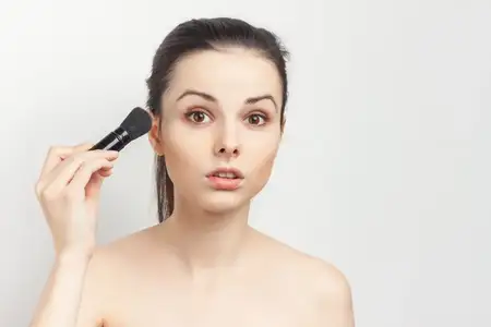 Женщина с обнаженными плечами, держащая в руках кисть и наносящая макияж