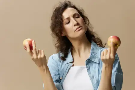 Задумчивая, сомневающаяся, кудрявая, красивая женщина в джинсах и повседневной рубашке смотрит на яблоко, позирует изолированно на бежевом пастельном фоне