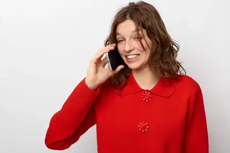 Улыбающаяся женщина в красном вязаном свитере прижимает смартфон к уху, выражая счастье и вовлеченность