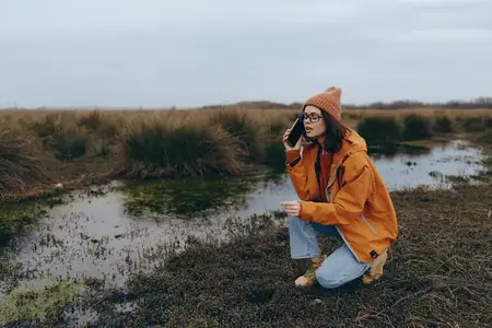 Отдых на природе: женщина в оранжевой куртке стоит на коленях на илистой земле у пруда среди травы и разговаривает по телефону