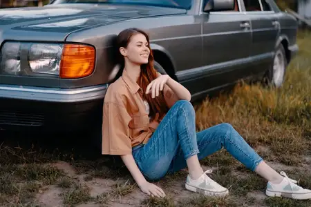 Счастливая молодая женщина в повседневной одежде сидит рядом с классическим автомобилем на травянистом поле, передавая радостную летнюю атмосферу, идеально подходящую для тем образа жизни и…