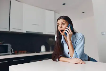 Женщина пользуется мобильным телефоном за кухонным столом, ведёт непринуждённый разговор в обстановке современного дома