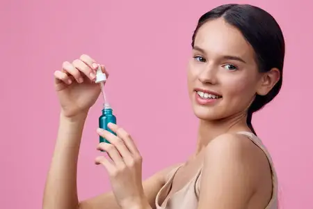 Улыбающаяся молодая женщина держит сыворотку для ухода за кожей, излучая радость и уверенность на фоне мягкого розового фона, пропагандируя красоту и благополучие
